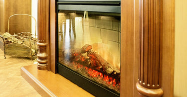 An electric fireplace features glowing artificial logs. It sits beside a metal basket in a warmly lit room.
